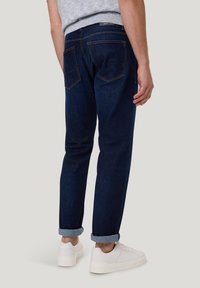 Man in donkerblauwe opgerolde jeans en witte sneakers, gezien van achteren tegen een effen lichtgrijze achtergrond.