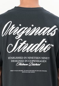 Black cotton t-shirt featuring large white text on the back. Text includes "Originals Studio™," establishment, design details, and branding.