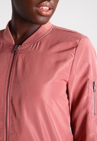 Pink bomber jacket with a smooth, shiny finish. Features a ribbed collar and front zipper, with a zippered pocket on the sleeve.