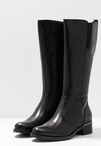 Un par de botas de cuero negras hasta la rodilla con tacones bajos y paneles elásticos laterales, de pie contra un fondo blanco liso.