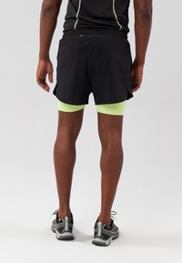 Pantaloni corti da corsa neri con fessure laterali, indossati sopra pantaloni aderenti verdi chiari. Scarpe da corsa grigie con dettagli neri. Dettagli riflettenti sulla maglietta.