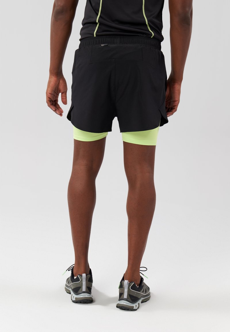 Pantalones cortos de running negros con aberturas laterales, llevados sobre pantalones cortos de compresión verde claro. Zapatillas de running grises con acentos negros. Detalles reflectantes en la camiseta.