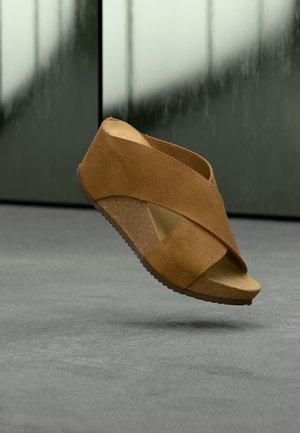 Smeđa antilop klinasta sandala s ukrštenim trakama i otvorenim prstima, lebdi iznad sivog poda na zamućenoj tamnozelenoj pozadini.