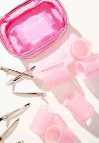 Estuche para accesorios de cabello rosa con un logo, que presenta varios rulos de cabello rosas y clips de cabello plateados dispuestos sobre un fondo claro.