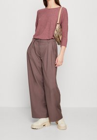 Top lungo a maniche lunghe di un delicato color malva abbinato a pantaloni a vita alta e gamba larga di colore marrone cioccolato. Scarpe beige con suola spessa e una borsa decorata completano il look.