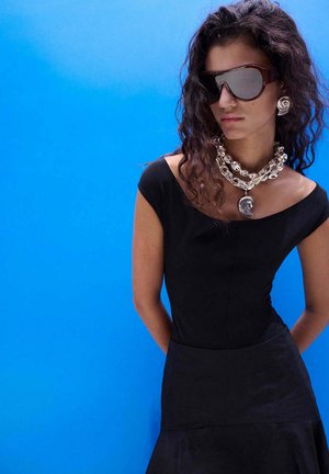 Woman with curly hair wearing oversized dark sunglasses, large silver jewelry, and a black off-shoulder dress against a bright blue background.