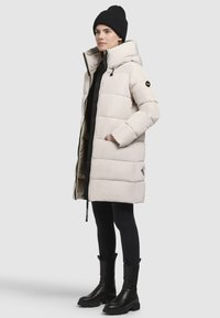 Lichtbeige pufferjas met een gewatteerd ontwerp, hoge hals en ritsluiting; gedragen over zwarte leggings en leren enkellaarzen. Zwart gebreide beanie.