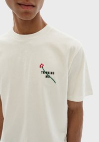 Witte katoenen t-shirt met een groene geborduurde steel met rode bessen en zwarte tekst "THINKING MU" op het linkerborstgebied.