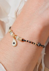 Bracelet perlé or et noir avec un pendentif en forme de goutte, présentant un centre blanc et une pierre noire. La base en fil tissé ajoute de la texture.