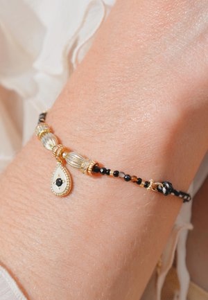 Bracelet perlé or et noir avec un pendentif en forme de goutte, présentant un centre blanc et une pierre noire. La base en fil tissé ajoute de la texture.