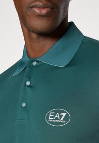 Tamsiai žalias polo marškinėliai su tekstūruotu paviršiumi, turintys klasikinį apykaklę, tris mygtukus ir išsiuvinėtą EA7 Emporio Armani logotipą.