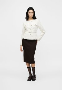 Fehér texturált dzseki nagy gombokkal és két elülső zsebbel, barna bordázott szoknyával és fekete zoknival, barna loaferekkel párosítva.