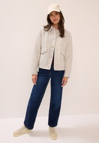 Kvinna som bär en krämfärgad fleecejacka, mörkblå jeans, krämfärgade sneakers och en krämfärgad keps, stående mot en ljus bakgrund.