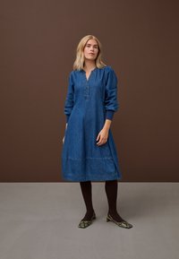 Femeie blondă stă în picioare purtând o rochie albastră din denim cu mâneci lungi, ciorapi opaci închiși la culoare și pantofi cu toc mic cu model de piele de șarpe pe un podea gri.