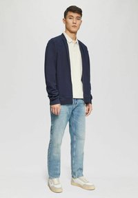 Marineblauer Reißverschluss-Sweatshirt, cremefarbener Poloshirt und hellblaue Jeans. Smart-Casual-Look mit weißen und beigen Sneakers. Einfaches Design, lockere Passform.
