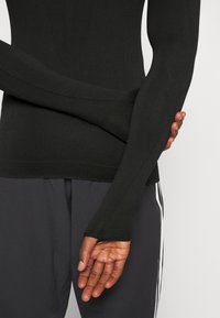 Schwarzes Langarmshirt mit taillierter Passform. Verfügt über eine glatte Textur, verlängerte Ärmel und einen engen Bund. Akzente umfassen Daumenschlitze.