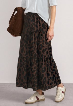 Person iført en hvid top, lang brun leopardprint nederdel, hvide sneakers med vinrøde striber og holder en brun ruskindstaske.
