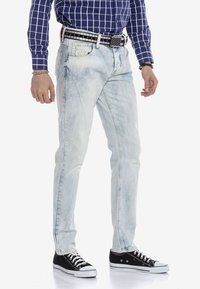 Hellblaue Denim-Jeans mit einem ausgewaschenen Finish, Slim Fit, fünf Taschen und gestickten Details. Getragen mit schwarzen Turnschuhen und einem gemusterten Gürtel.
