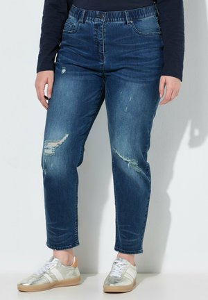 Person iført slidte blå jeans, mørk langærmet skjorte og sølvfarvede sneakers, der står op ad en hvid væg.