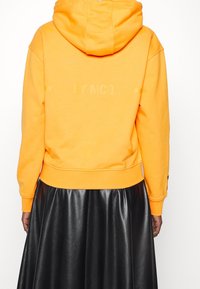 Sudadera con capucha naranja y bolsillo canguro, con un logo en la parte posterior. Combinada con una falda corta de cuero negro brillante.