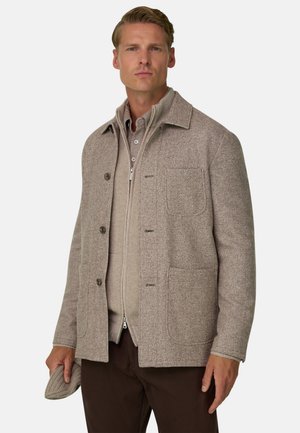 Un homme porte une veste texturée taupe boutonnée sur le devant, par-dessus un pull en maille zippé, et tient une casquette assortie devant un fond blanc uni.