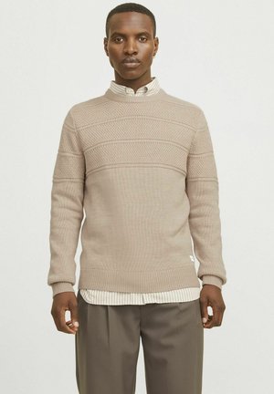 Pull en tricot beige avec des rayures horizontales texturées, col rond, poignets et ourlet côtelés, superposé à une chemise rayée blanche et grise.