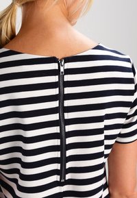 Bakifrån av en person som bär en svart och vit horisontell randig topp med en dragkedja som går ner längs mitt bak på övre delen.