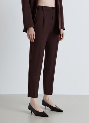 Donna che indossa pantaloni alla caviglia su misura color marrone scuro abbinati a scarpe nere a punta con tacco basso, in piedi davanti a uno sfondo semplice.