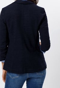 Blazer texturé bleu marine avec un design sur mesure, fermeture à un bouton, et manches ajustées, porté sur une chemise bleu clair avec un jean bleu.