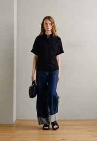 Svart kortärmad skjorta med knappar, mörk denim vidbyxor med kontrasterande ärmslut, svart handväska och svarta platta sandaler. Enfärgad bakgrund.