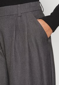 Grijze geplooide broek van een gestructureerde stof, met een zijzak en een platte tailleband. De kleuren zijn gedempt, geschikt voor formele kleding.
