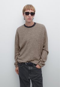 Beige knit sweater with long sleeves and a round neckline, paired with dark gray jeans. The individual wears black sunglasses and a dark belt.