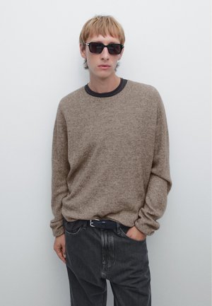 Beige stickad tröja med långa ärmar och rund halsringning, kombinerad med mörkgrå jeans. Personen bär svarta solglasögon och ett mörkt bälte.