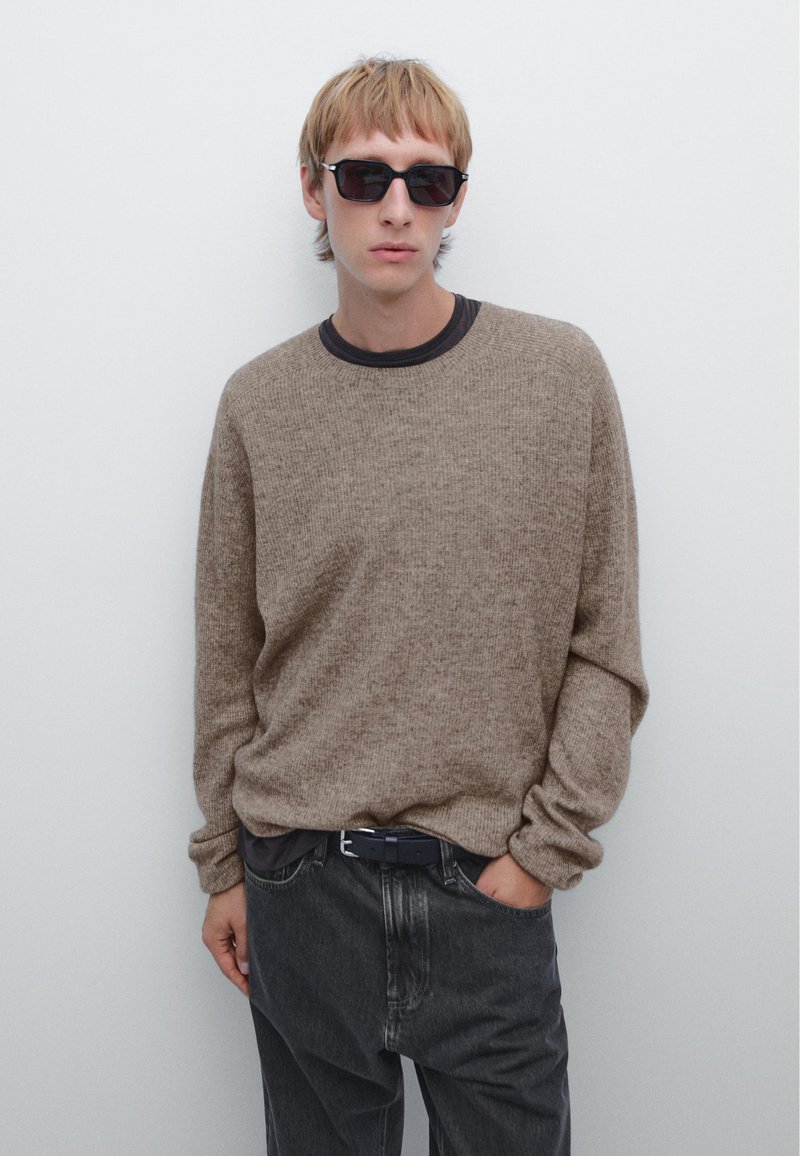 Beige knit sweater with long sleeves and a round neckline, paired with dark gray jeans. The individual wears black sunglasses and a dark belt.