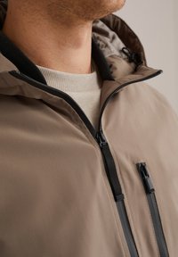 Chaqueta marrón con cremallera y capucha, con cremalleras y acentos negros. Fabricada en un tejido suave, incluye un alto cuello y un diseño elegante.