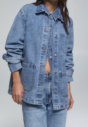 Person iført lagdelt lyseblå denimjakke og jeans med opkørte ærmer og let blottet mave mod ensfarvet baggrund.