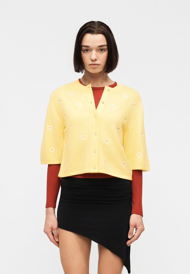 Femme portant un cardigan jaune à manches courtes avec des fleurs blanches par-dessus un haut rouge à manches longues et une jupe noire asymétrique, debout devant un fond blanc.