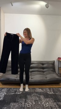Femme en débardeur bleu marine et leggings noirs debout pieds nus dans un salon, tenant un pantalon noir devant un canapé gris.