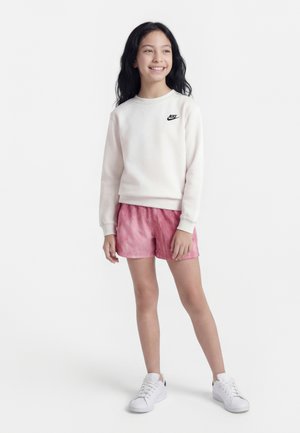 Dziewczyna stoi, ubrana w białą bluzę Nike, różowe szorty i białe trampki, uśmiecha się i patrzy lekko na bok.