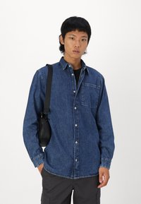 Camisa de mezclilla en azul oscuro con diseño abotonado, bolsillo en el pecho y un corte relajado. Llevada con pantalones cargo grises y un bolso de hombro negro.