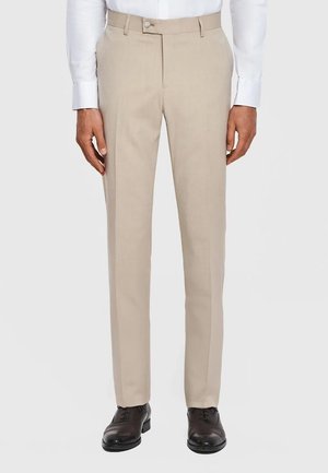 Mężczyzna ubrany w beżowe formalne spodnie, czarne eleganckie buty i białą koszulę z długim rękawem, stoi z rękami swobodnie opuszczonymi wzdłuż ciała.