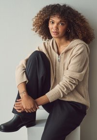 Felpa beige con zip e maniche a coste, indossata con pantaloni neri larghi. Stivaletti neri visibili. Soggetto seduto contro un muro chiaro.