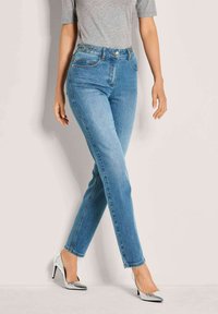 Frau in hellblauen, eng anliegenden Jeans und silbernen High Heels, die mit verschränkten Beinen vor einem neutralen Hintergrund steht.