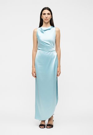 Femme aux longs cheveux foncés portant une robe de soirée en satin bleu clair sans manches et des escarpins noirs ouverts sur un fond uni.