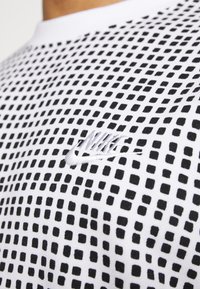 Camiseta atlética blanca con un diseño estampado de pequeños cuadrados negros, con un logo de Nike bordado en negro en el pecho.