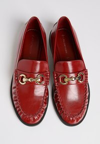 Red leather loafers with a rounded toe, featuring a gold chain accent and stitched detailing along the edges. Smooth surface texture.