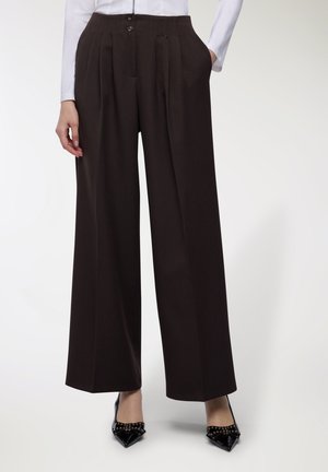 Donna che indossa pantaloni larghi a vita alta di colore marrone scuro con plissettature, abbinati a tacchi a spillo neri con borchie e a una maglietta bianca a maniche lunghe.