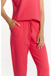 Różowy zestaw odzieży domowej składający się z luźno dopasowanej bluzki oraz spodni z elastycznym pasem i sznurkiem z złotymi końcówkami; gładka faktura materiału.