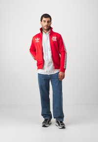 Red zip-up jacket with black and white stripes, logo on chest, worn over a white striped shirt and blue jeans, paired with sporty sneakers.