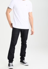 Vit bomullströja med korta ärmar, mörka denimjeans och svarta kanvas sneakers med vita snören, stående mot en neutral bakgrund.